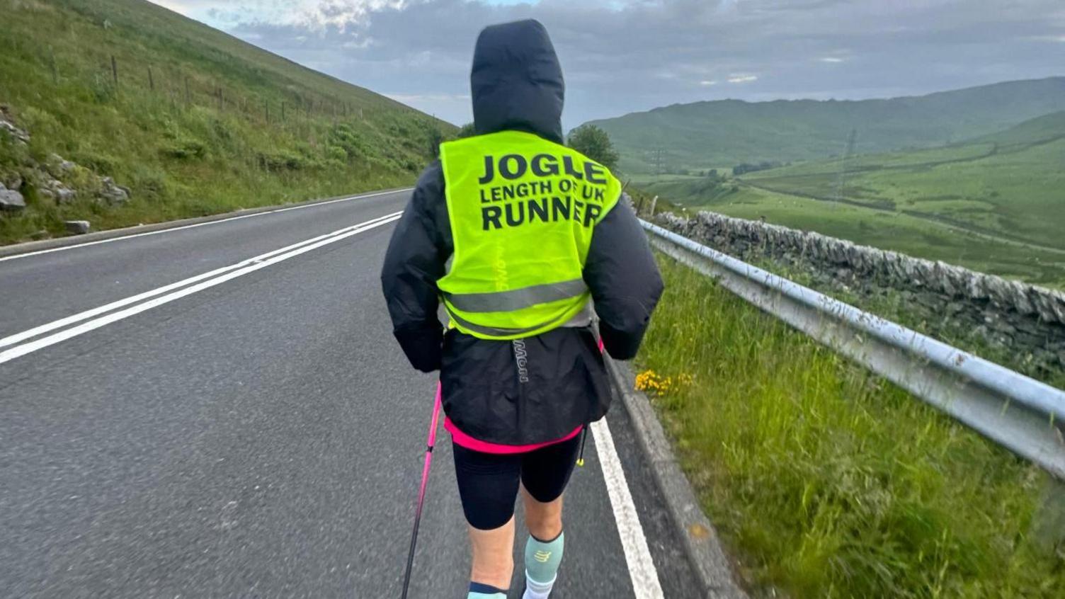 The back of a man running on a main road wearing a bright yellow jacket and black shorts.