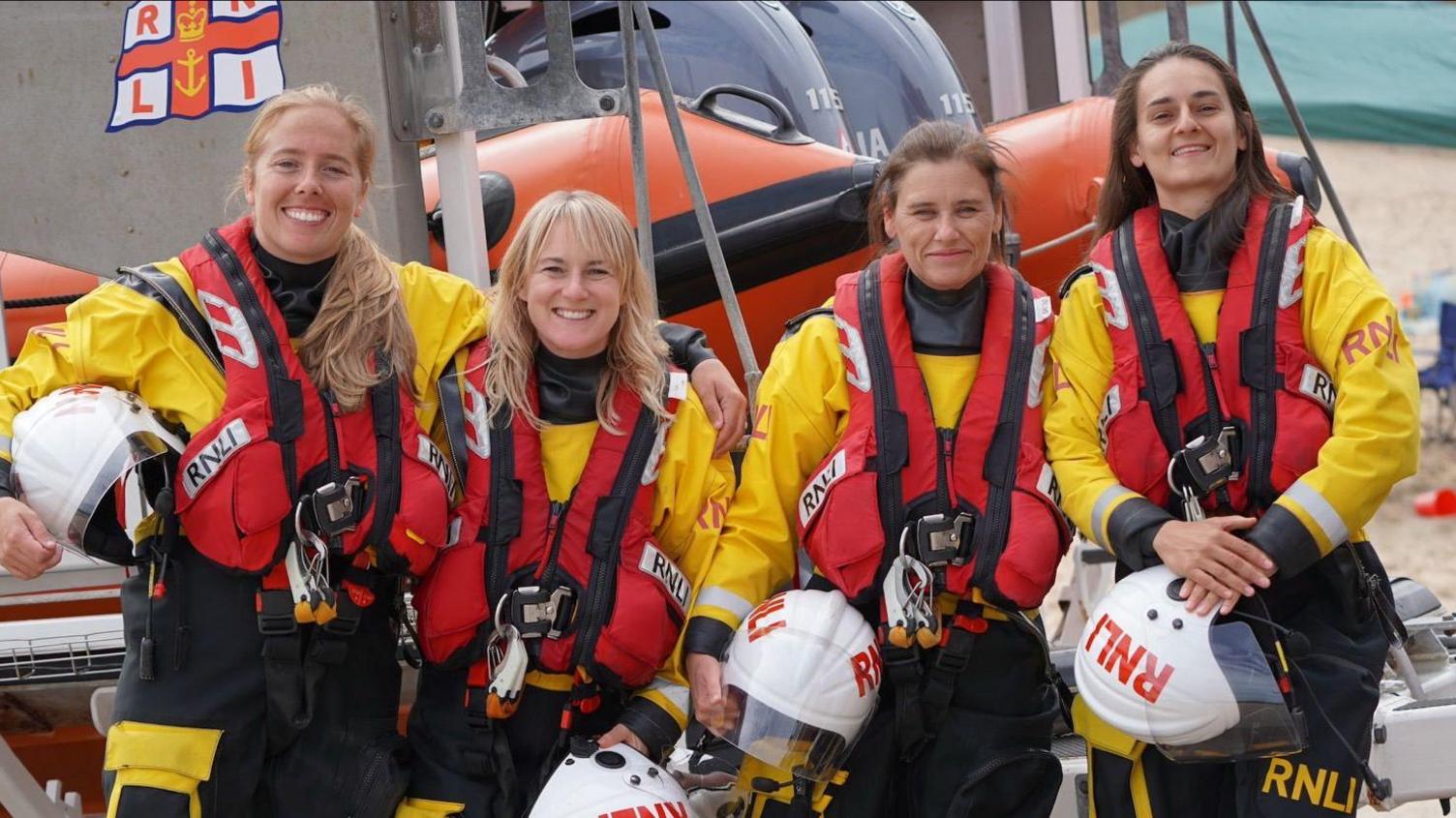 RNLI crew members Anna Heslop-Latif, Sarah Whitelaw, Karen Pearce and Jane Heslop-Latif. The women are wearing yellow and black uniforms and red lifejackets. They stand in front of a RIB outside the Cullercoats station.
