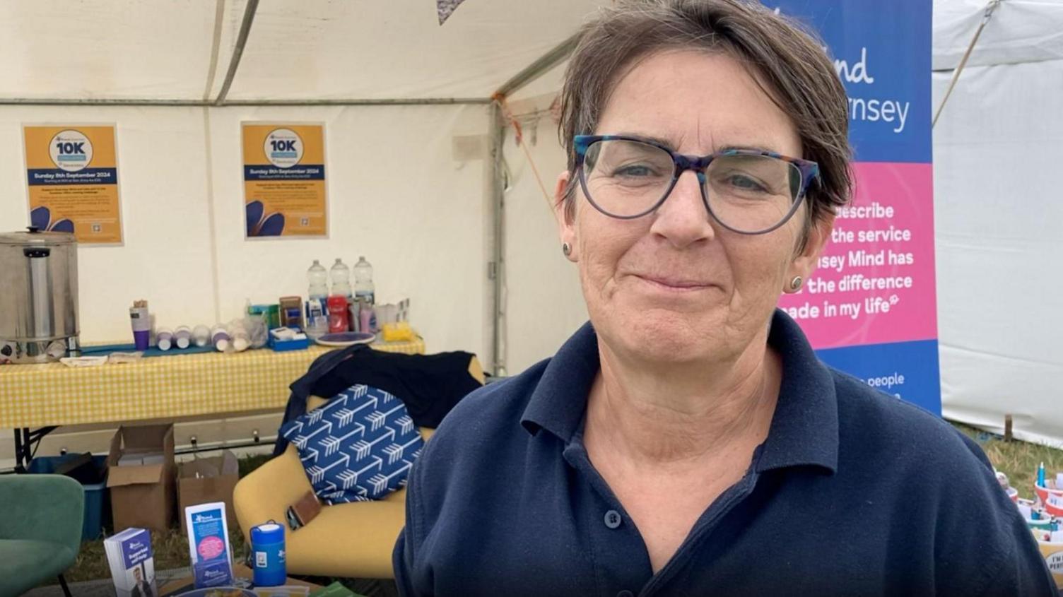 Amanda Hibbs - who has short brown hair, tortoiseshell blue rimmed glasses - is pictured smiling while wearing a navy polo top. Behind her are some chairs and a long table with a tea urn and cups in a white tent