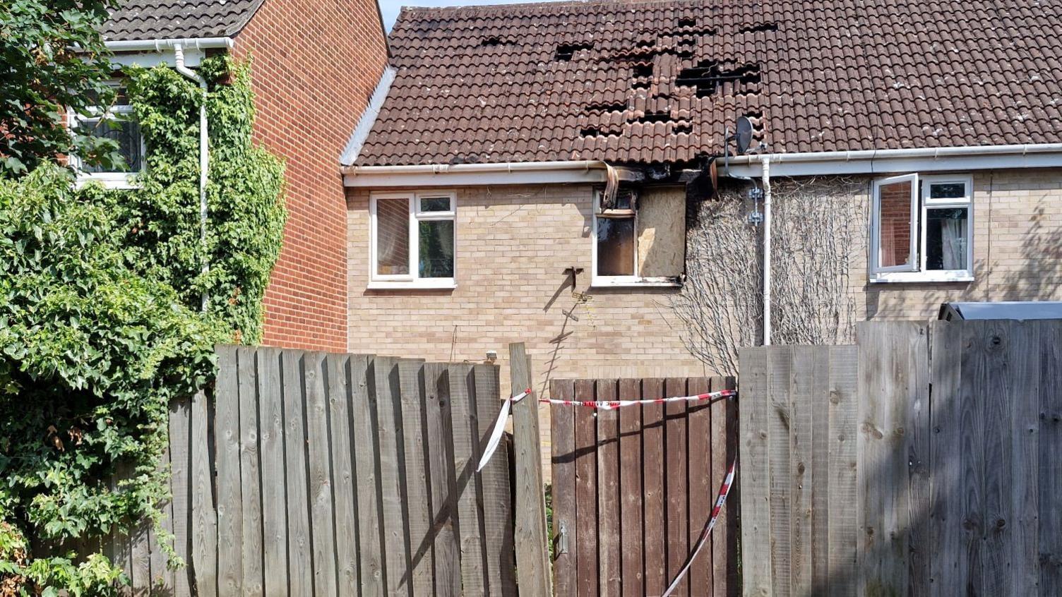 Smoke damage to the roof and a first-floor window of a small, two-storey semi-detached house. There is still some emergency services tape on the back gate, and some roof tiles seem to be missing.