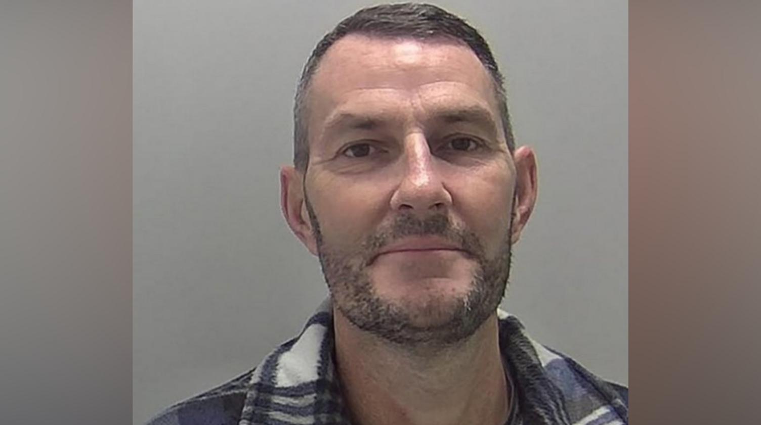 A man with dark hair and a beard in a blue and white checked shirt
