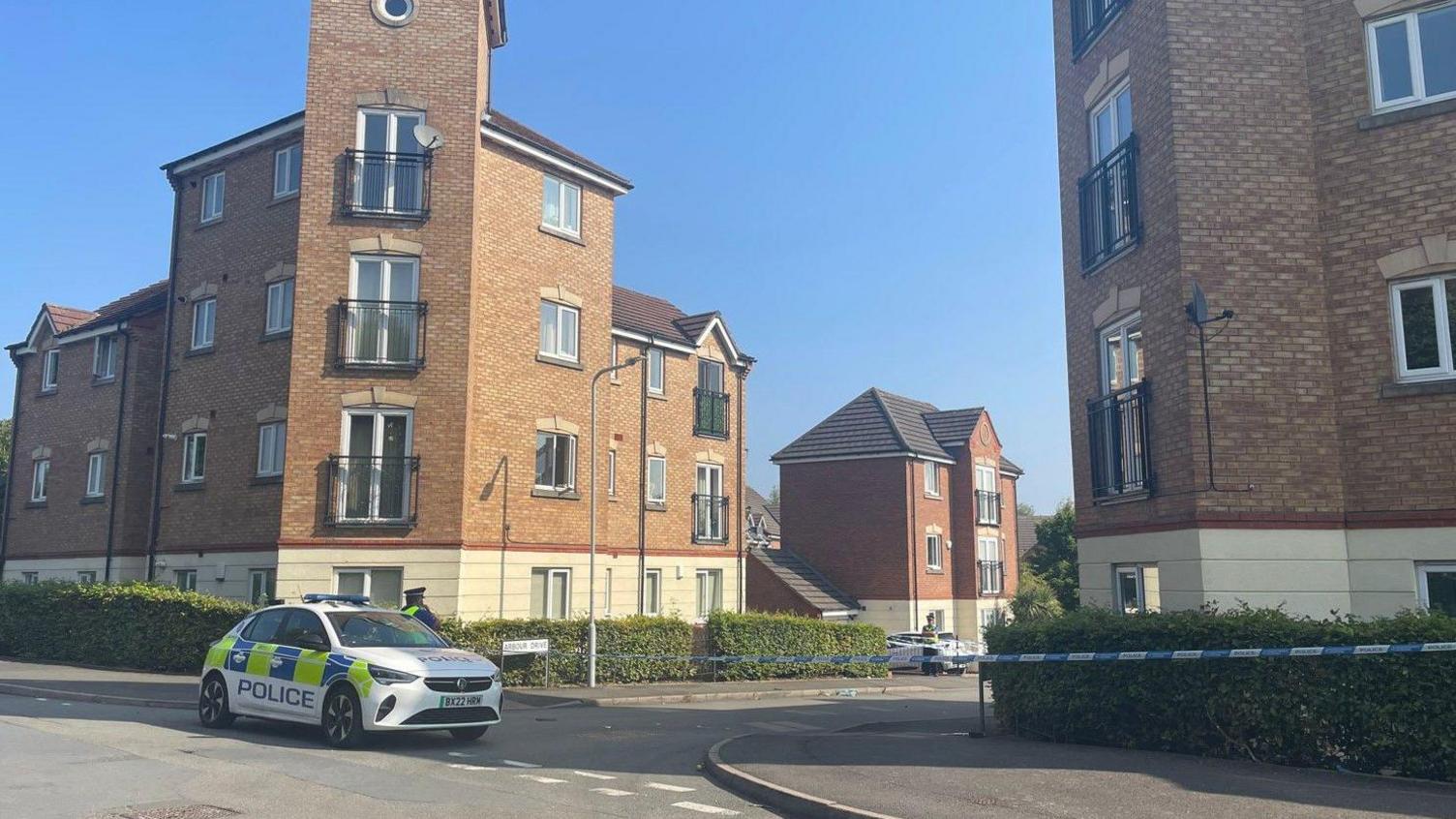 A police car is parked at a road junction near to blue and white police tape. There are buildings from left to right on the photo.