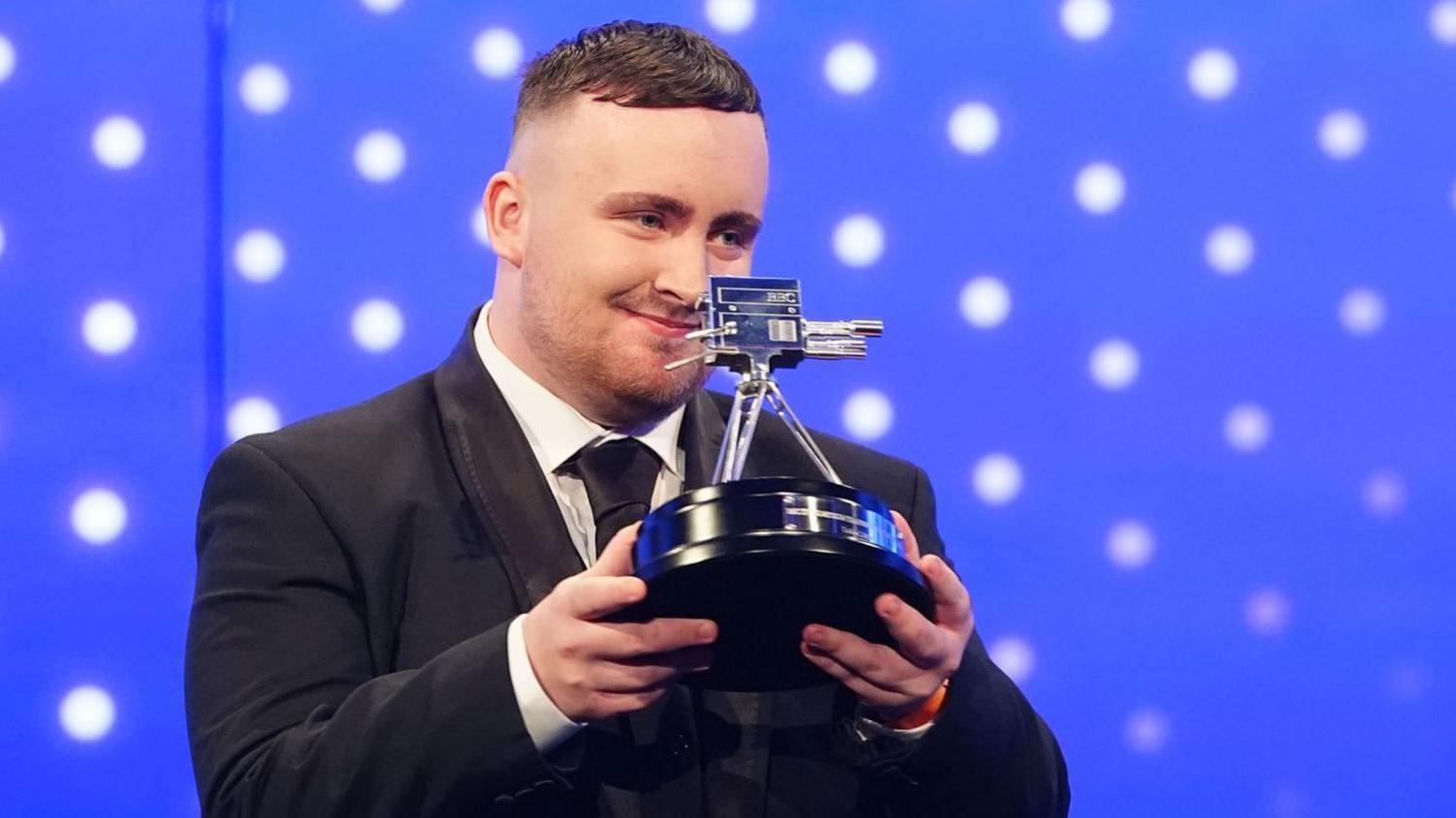 Luke Littler, dressed in a black suit with a white shirt and a black tie, holds up the BBC Young Sports Personality of the Year trophy.