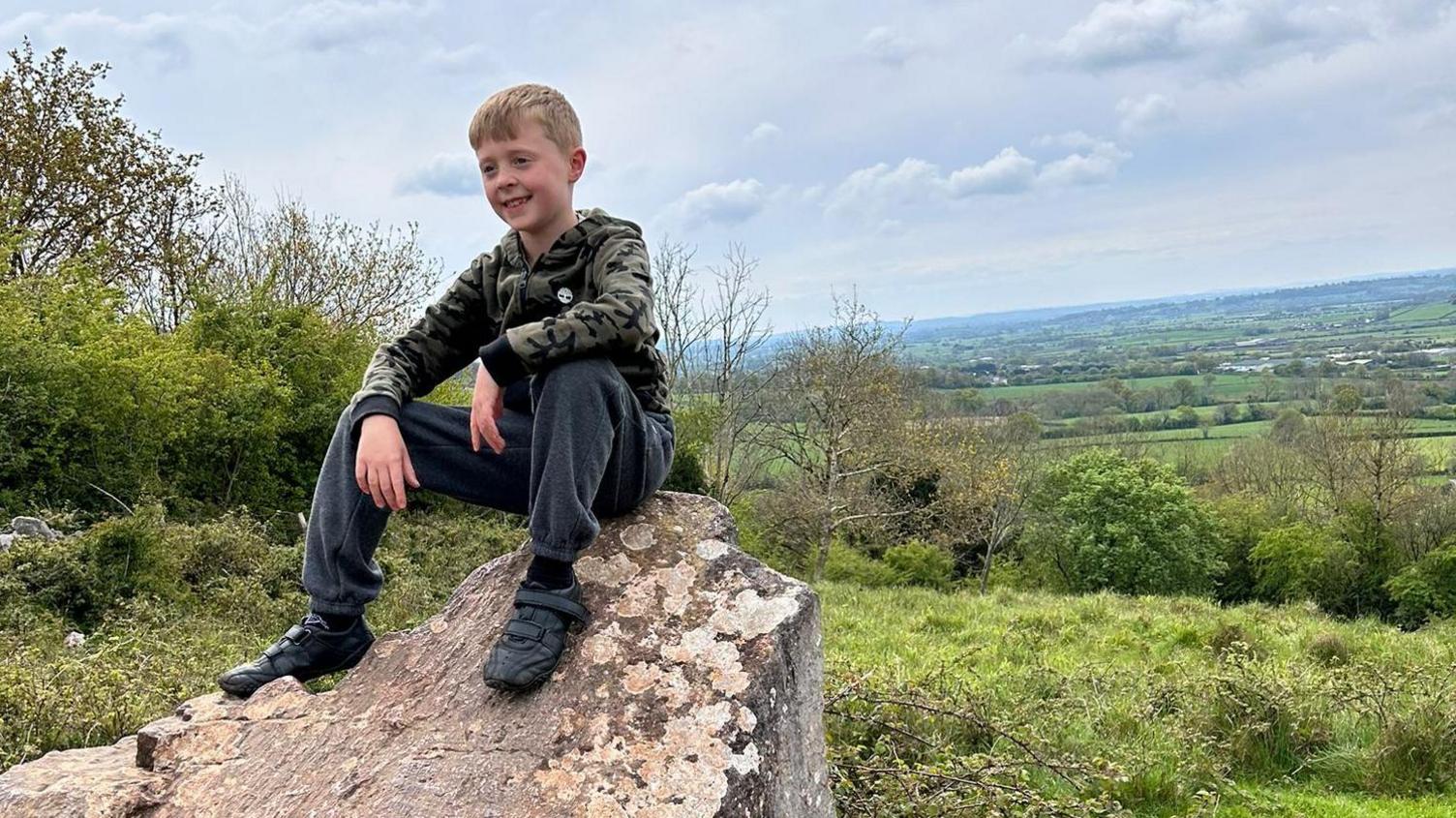 A young boy in a tracksuit sitting on a rock with a grassy view behind him.