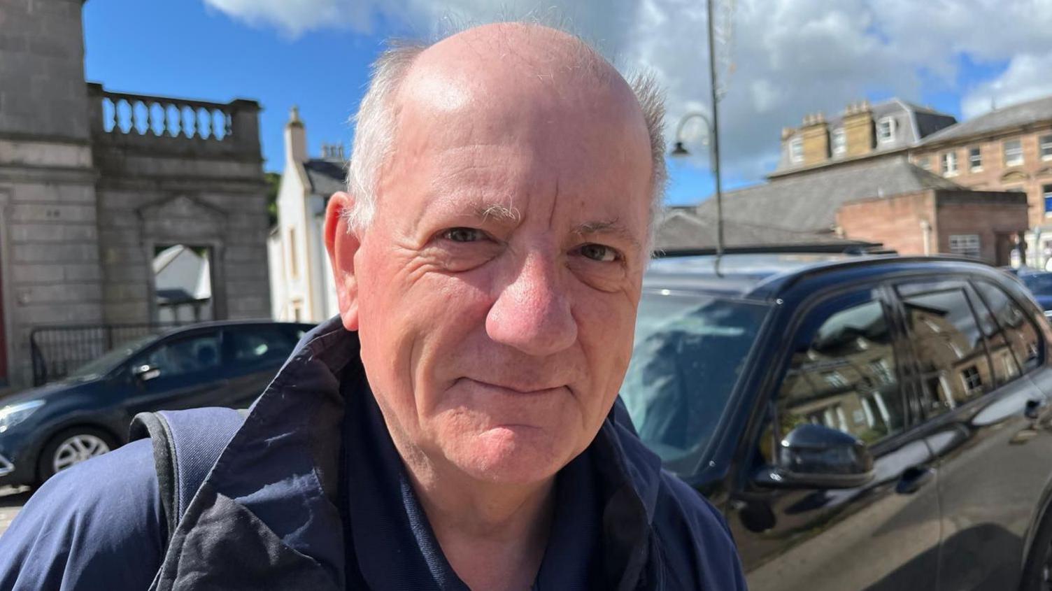 Bruce is standing outside in Dingwall. He has grey hair and is smiling. He is wearing a blue jacket and strap of a rucksack is visible on one shoulder. There are cars parked on the street behind him.