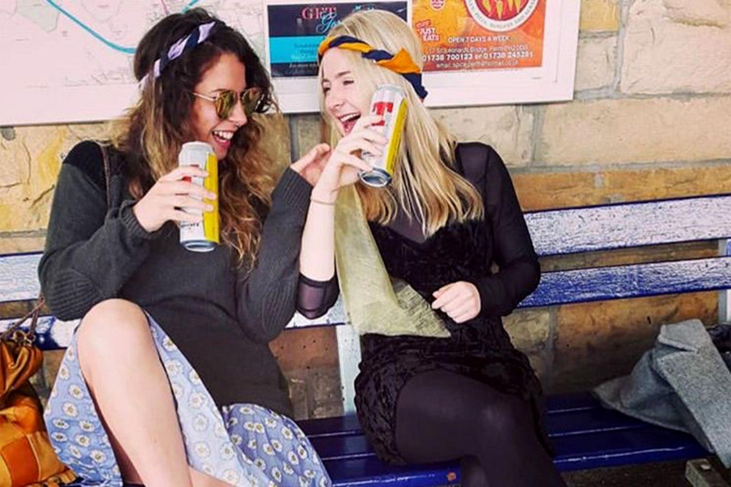 Hazel laughing with a friend - they are both sitting on a blue bench and holding cans of lager 