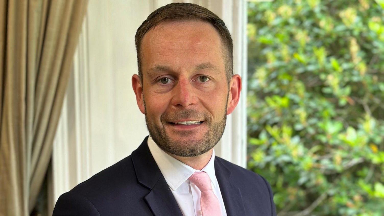 Dan Ainsworth smiles at the camera, wearing a blue blazer, white shirt and pink tie. 