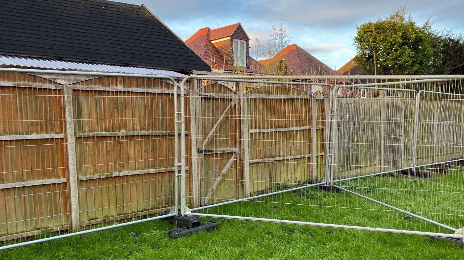 Heras fencing on the edge of a field running parallel to a wooden garden fence. There is a gate in the middle of the wooden fence which is now unable to open. 