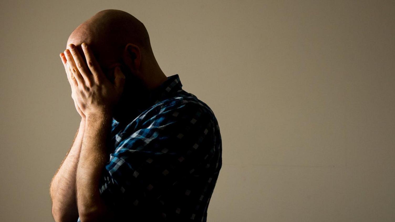 Head and shoulder shot of a standing man wearing a blue checked shirt and clasping his hands to his face in a manner suggestive of despair 