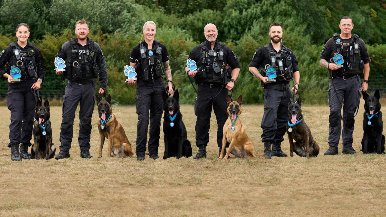 Two women and four men are standing in a line outside in police uniform, all holding their blue paw trophies. Their dogs are next to them, all wearing their blue paw medals. 