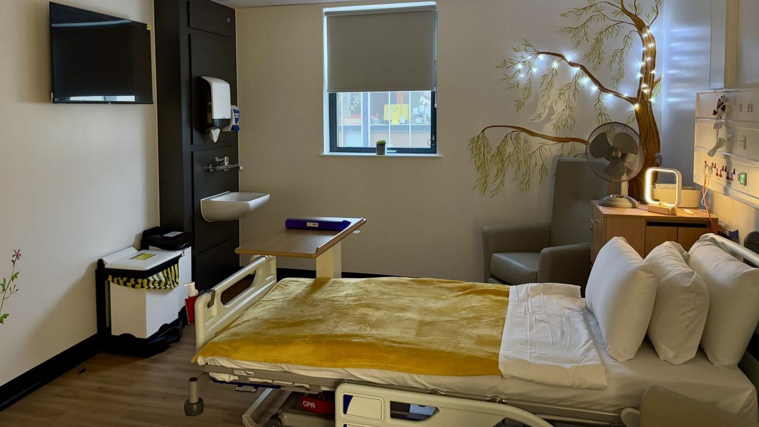 Inside a bedroom at the new palliative care unit. There is a hospital bed with a golden throw on top. Surrounding the bed is a bedside unit, bins and sink. On the wall is the painting of a tree with lights across the branches