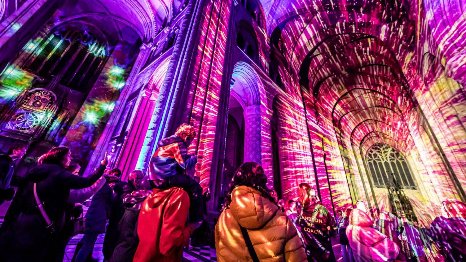 A light installation, with vivid purple and yellow colours, is lighting up the inside of a cathedral building. There are people in the foreground, who are also lit up by the display, with some taking photos on their phones.