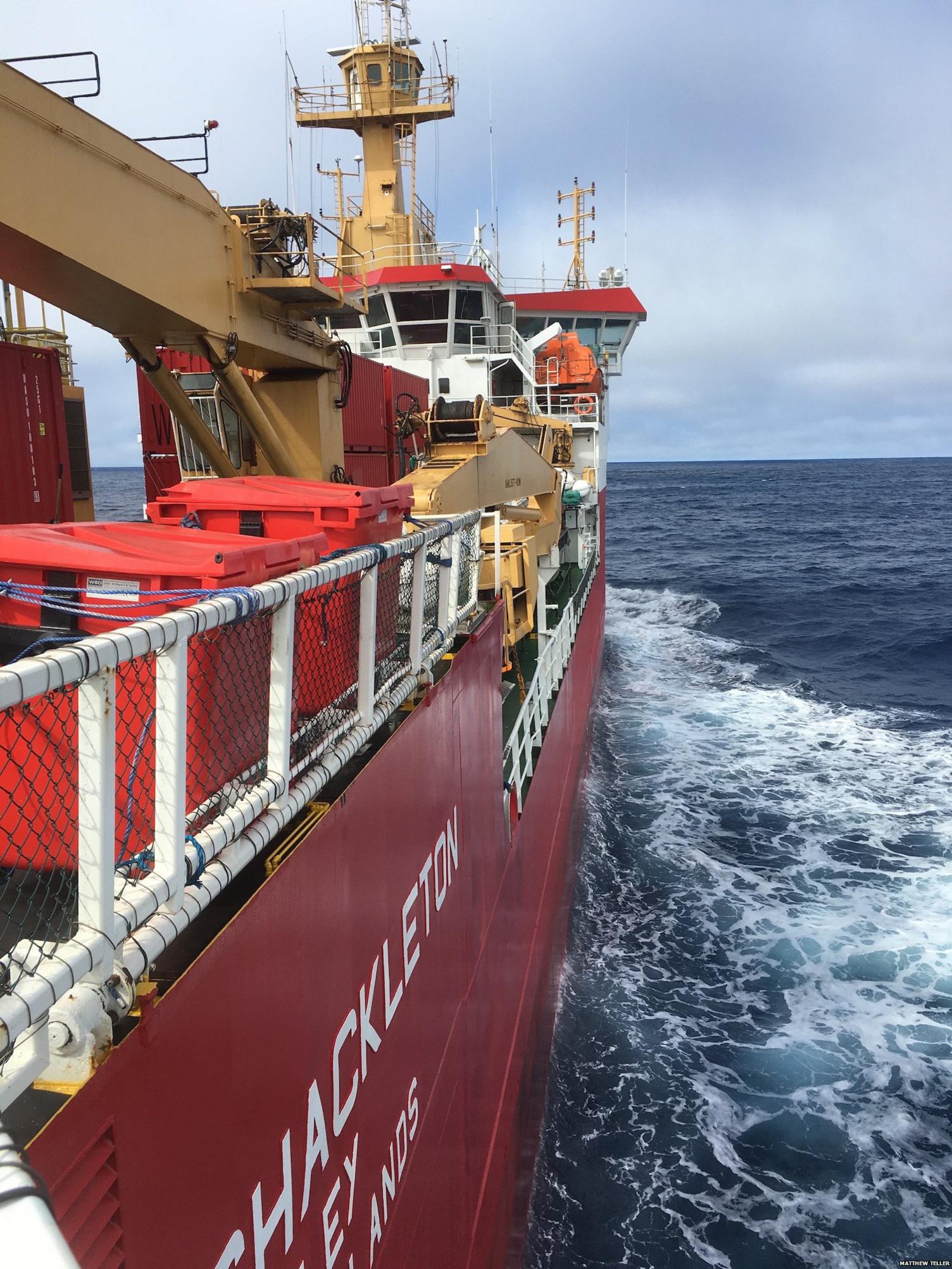 View of the back of a bright red ship ripping through the ocean. Grey skies in the background.