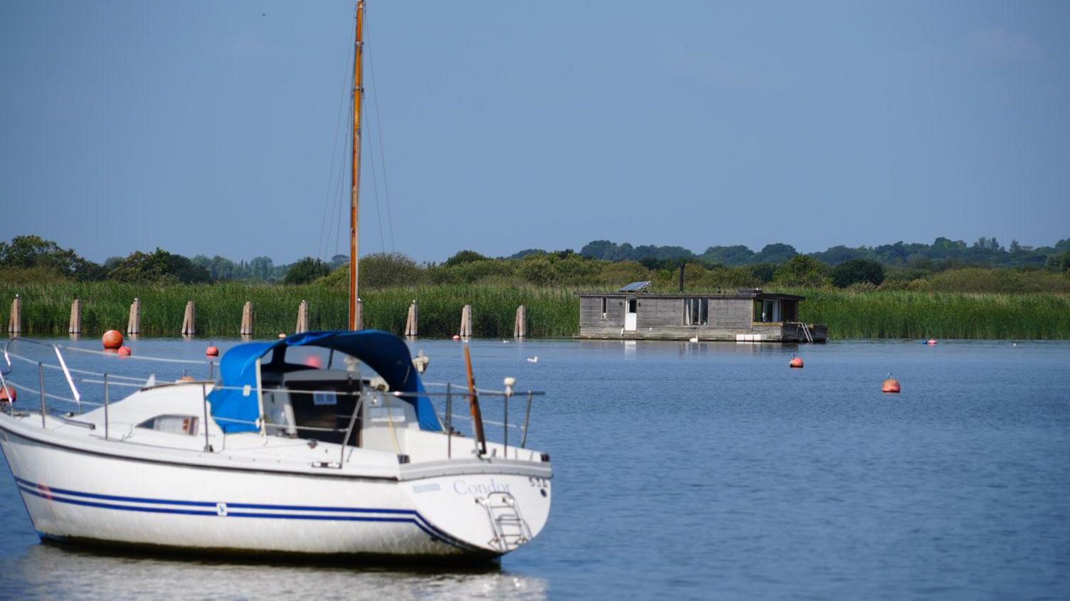 A view of Oulton Broad. A yacht rests in the water with a houseboat in the distance.