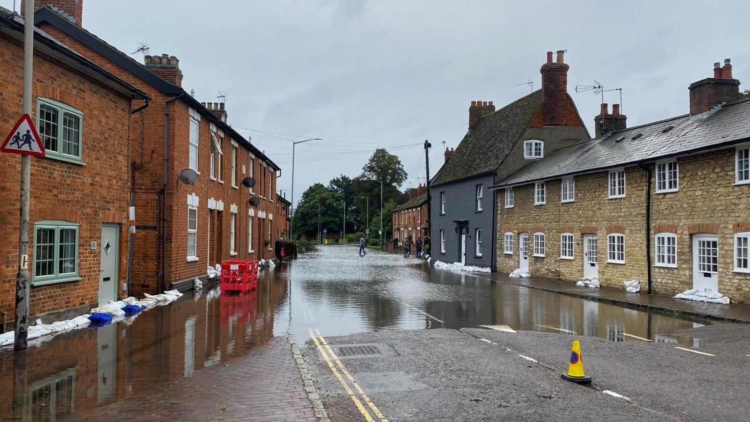 A road lined by cottage style houses with part of the road flooded, and houses lined by sand bags.