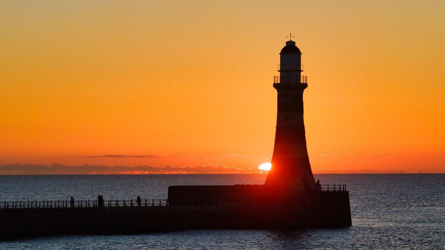 The sun rising behind Roker lighthouse which is just a silhouette in the light. The sky is bright orange and the sea surrounding the the pier in lighthouse is dark blue and calm.