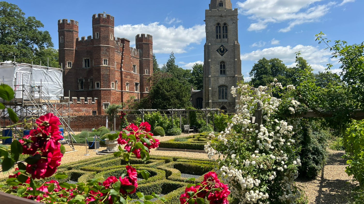 The Queen Katherine Knot Garden is full of flowers with red and white ones in the foreground.
Behind them are two symmetrical mazes - made from short bushes. Stone lines a path around the garden and there are trees and shrubbery around the edges. In the background the red bricked Buckden Towers can be seen to the left of a tall church tower which has a clock in the centre.
