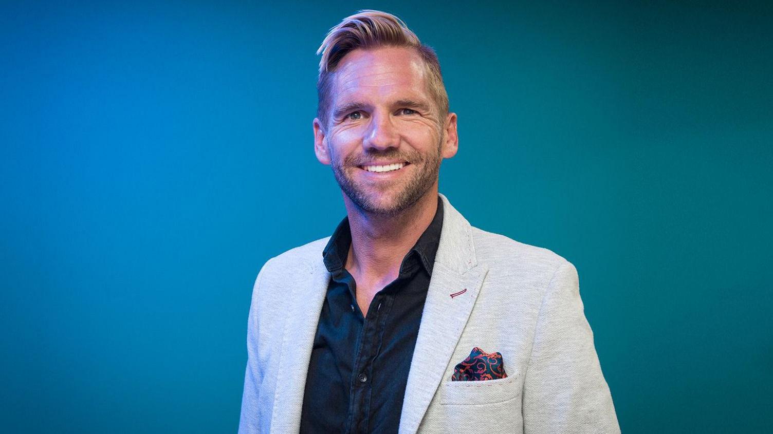 Jake Moore, who is smiling and has blonde hair and a beard. He is wearing a light grey jacket with pocket square over a navy blue open-necked shirt. He is set against a bright blue background.