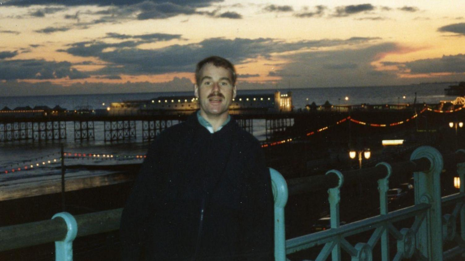 A man stands with a beach and a pier behind him as the sun sets. 