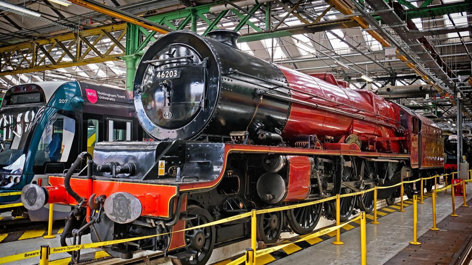A red vintage steam locomotive on display in a factory setting, surrounded by yellow caution barriers