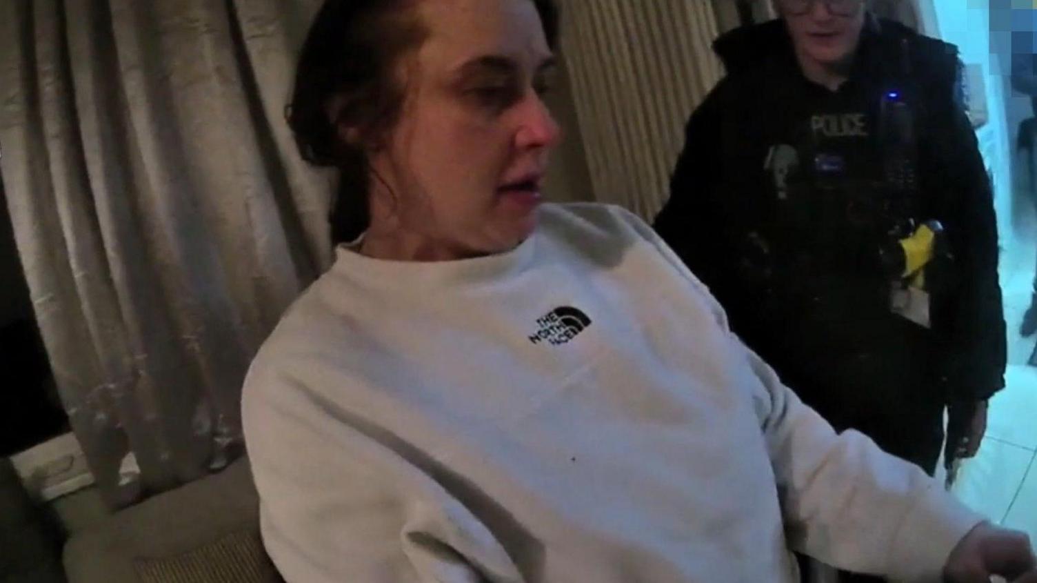 A still of a bodycam video showing a woman wearing a white jumper with "The North Face" on it being arrested. A police officer with glasses is behind her.