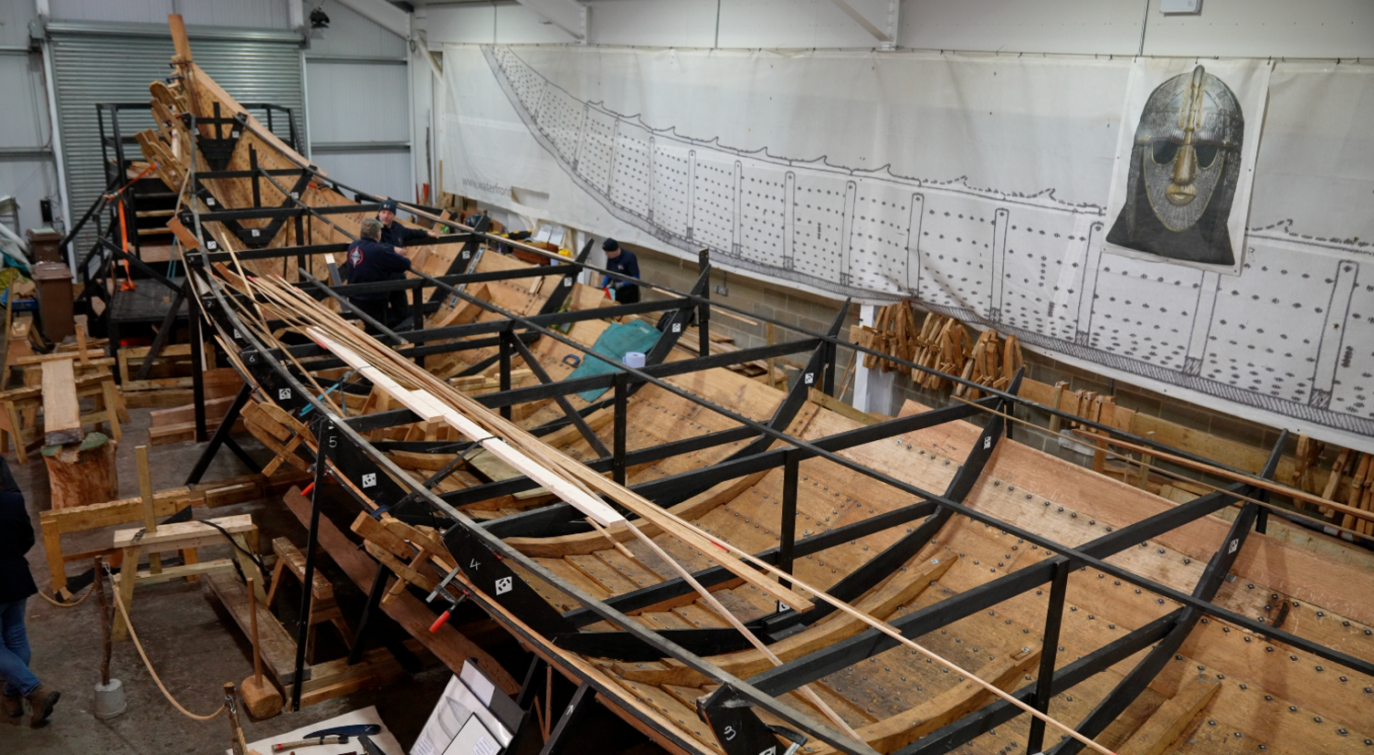 A view of the longship in the process of being built inside a large warehouse. Planks of wood lie either side of the ship. There is an image of a silver helmet above the ship. 