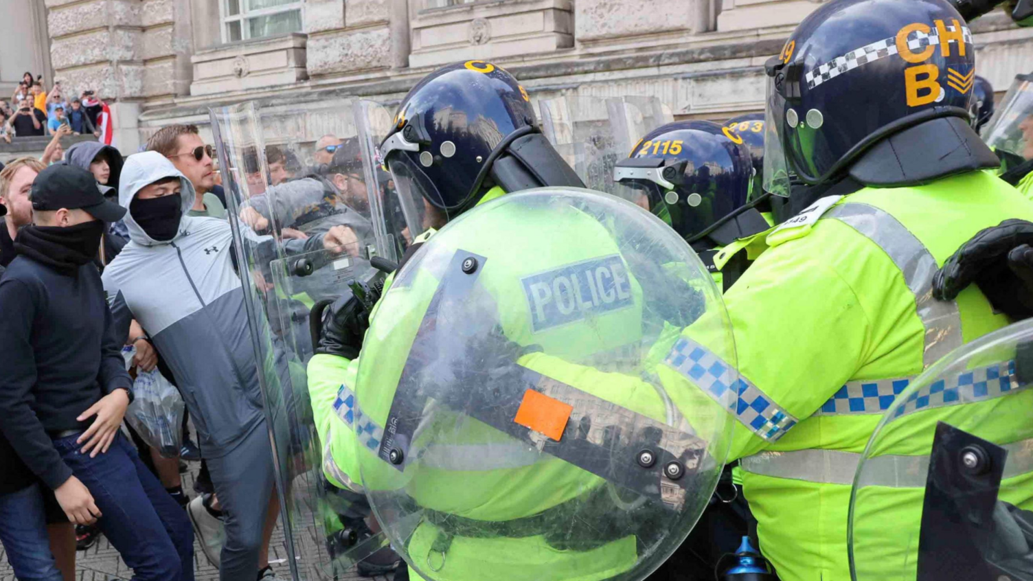 Police in yellow high vis , wearing black helmets and carrying clear riot shields push back against a crowd of protesters - one at the front in a grey tracksuit kicks at a shield.