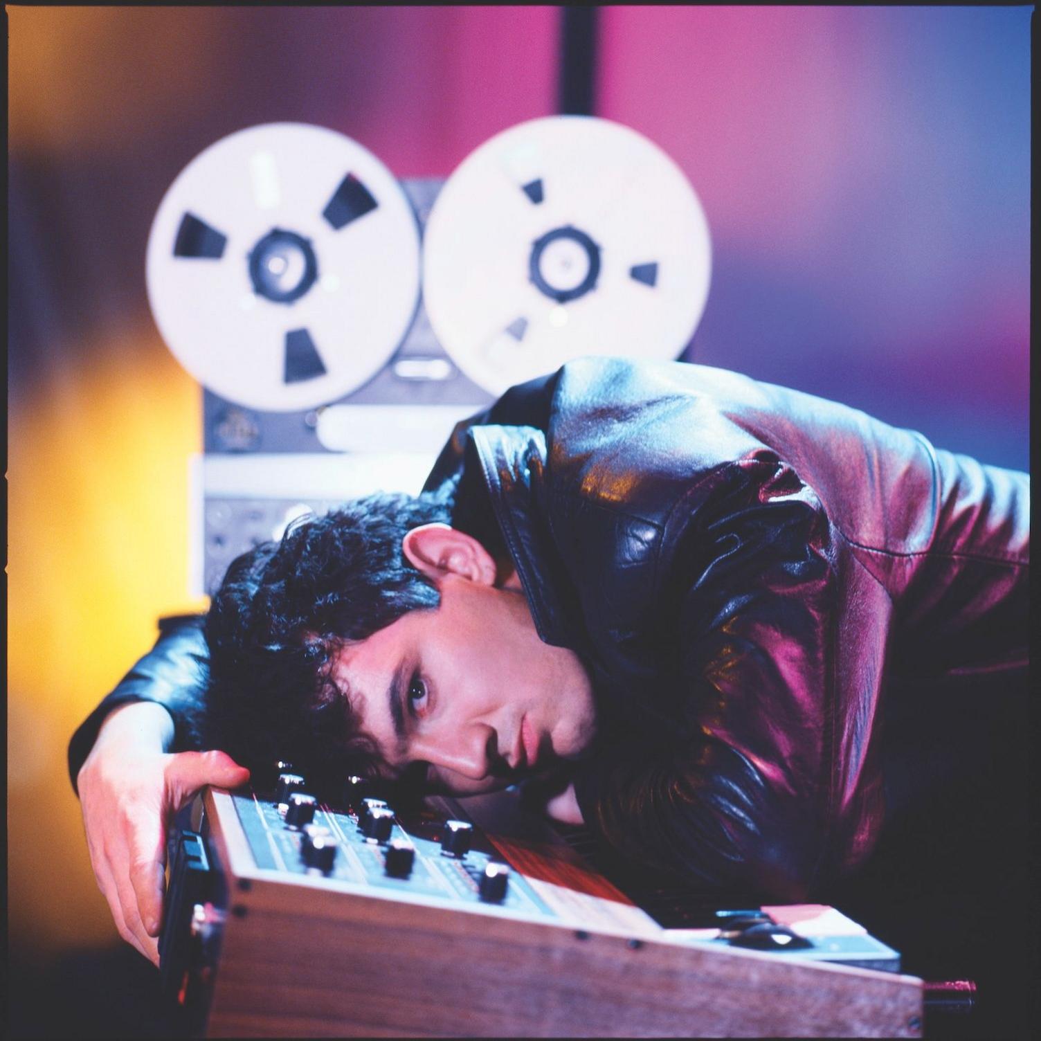 Dave Ball is seen lying on top of a 1980s synthesizer, with a reel-to-reel tape machine in the background