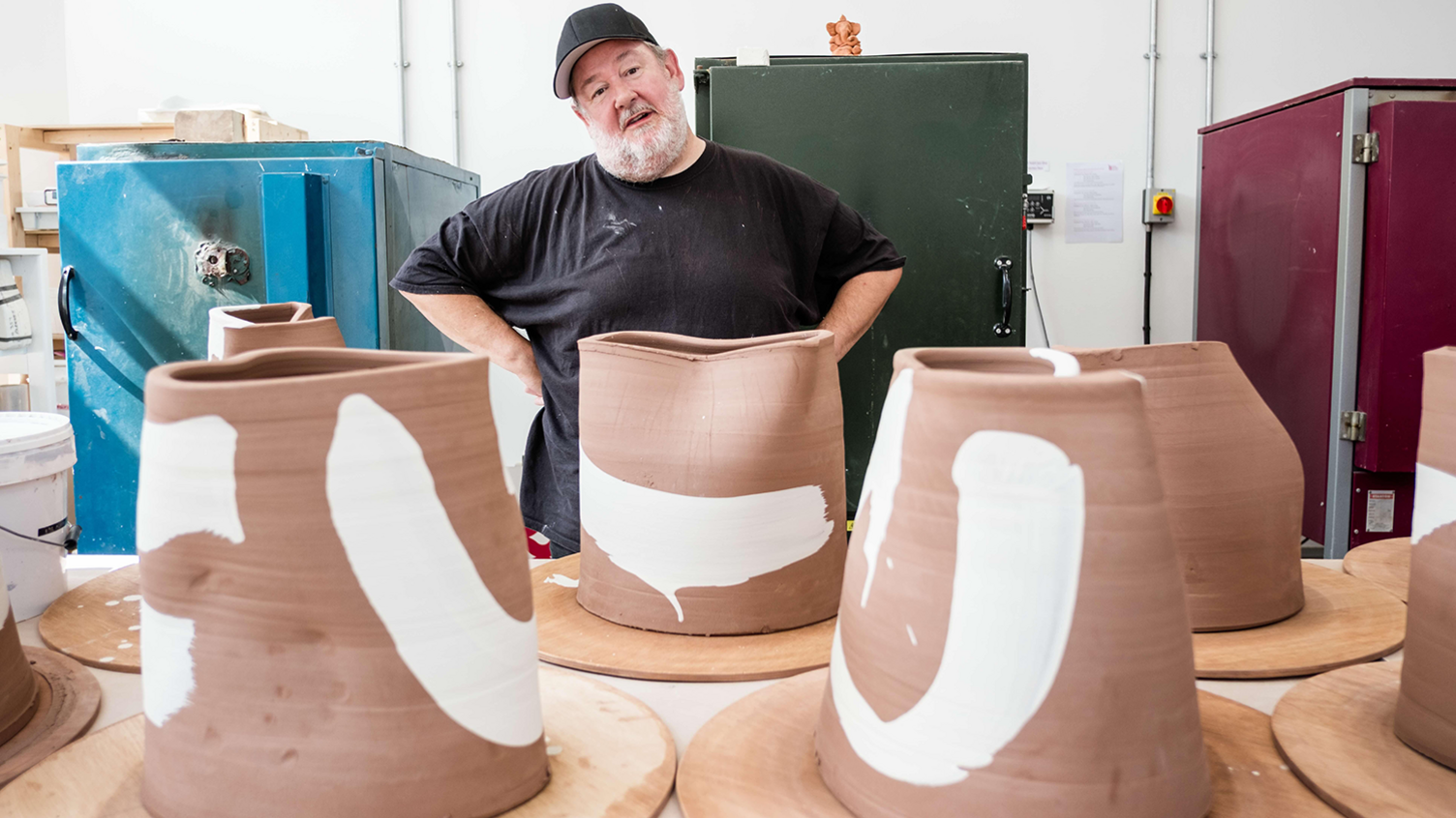 Johnny Vegas stands in a workshop with his hands on his hips and head cocked to one side. He is standing behind a table with large brown clay models with white paint on them.
