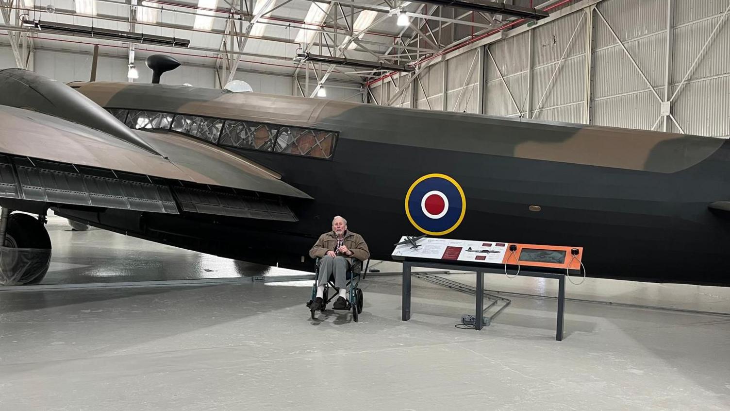 Donald Merryweather sitting in front of a Wellington bomber at the museum. He is in a wheelchair next to an information board about the aircraft, which bears the RAF's red, white and blue roundel.