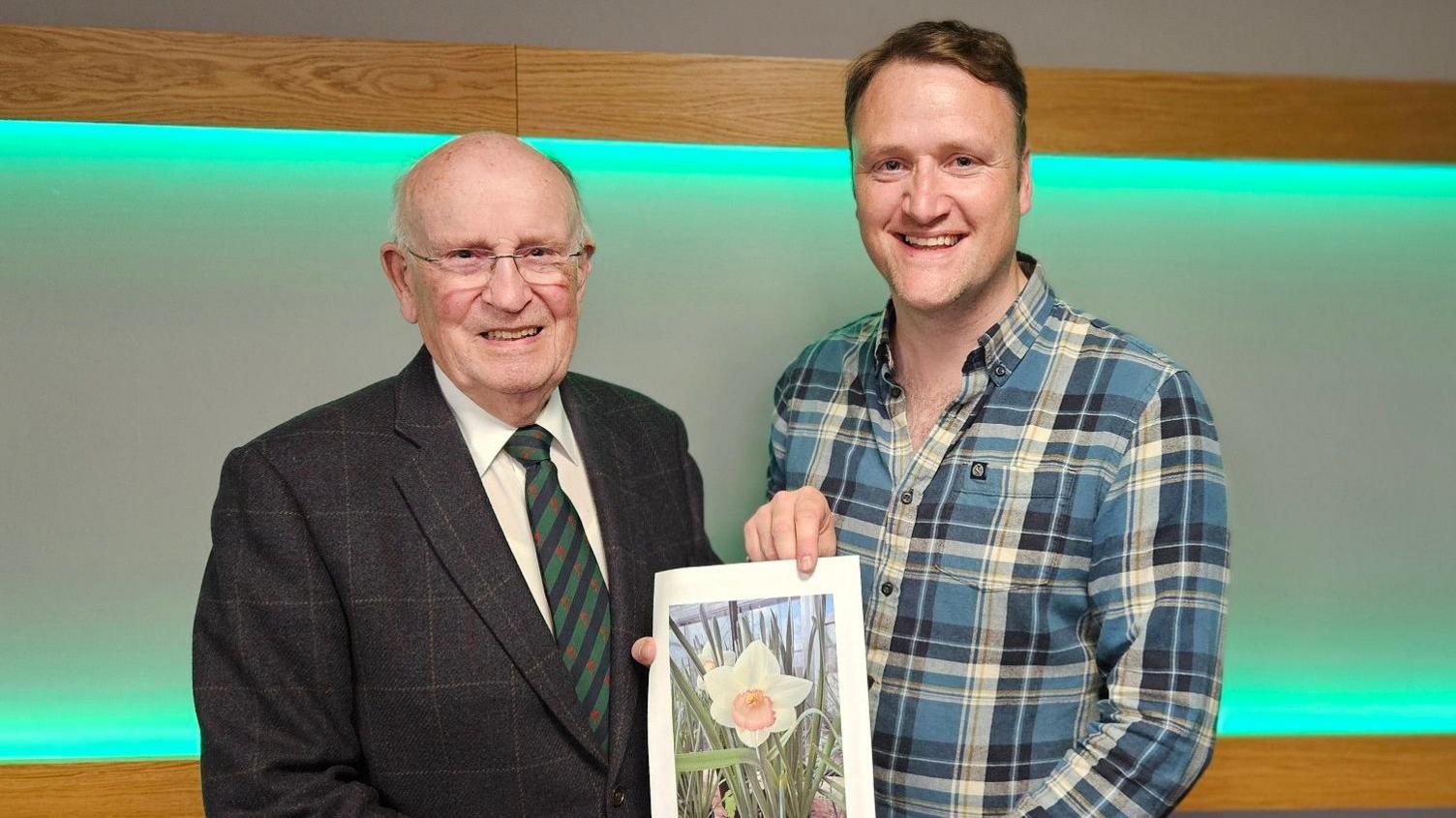 An elderly man in a dark suit and green striped tie stands to the left of another man in a chequered blue shirt. They are both holding a photograph of a daffodil.