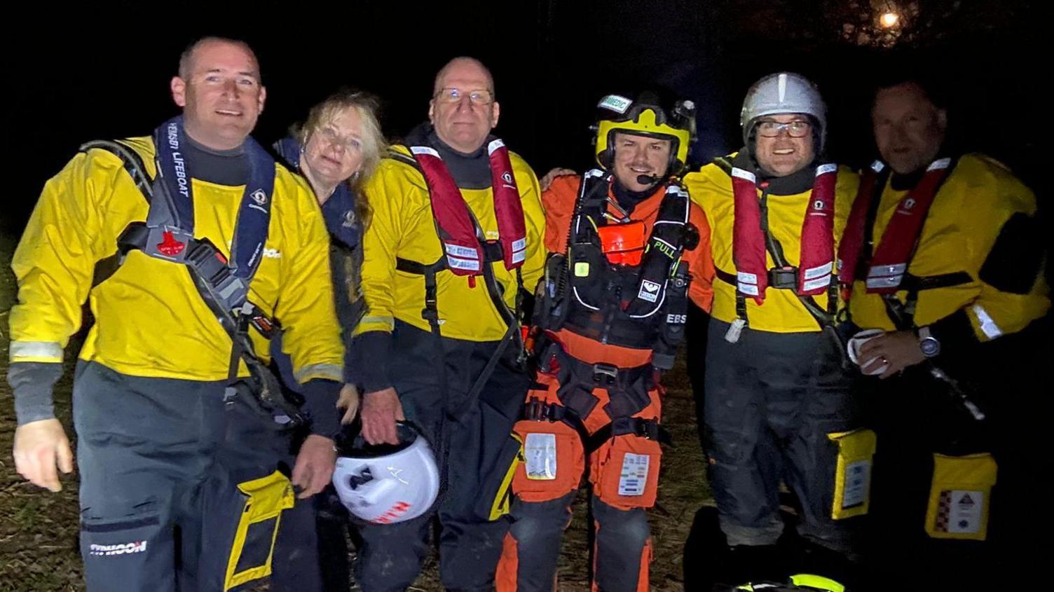 Six people standing in a line, in the dark, outside. Five are wearing lifeboat uniforms, including yellow tops and blue trousers, and one helicopter paramedic in the middle, in an orange jumpsuit, with a  helmet on. 