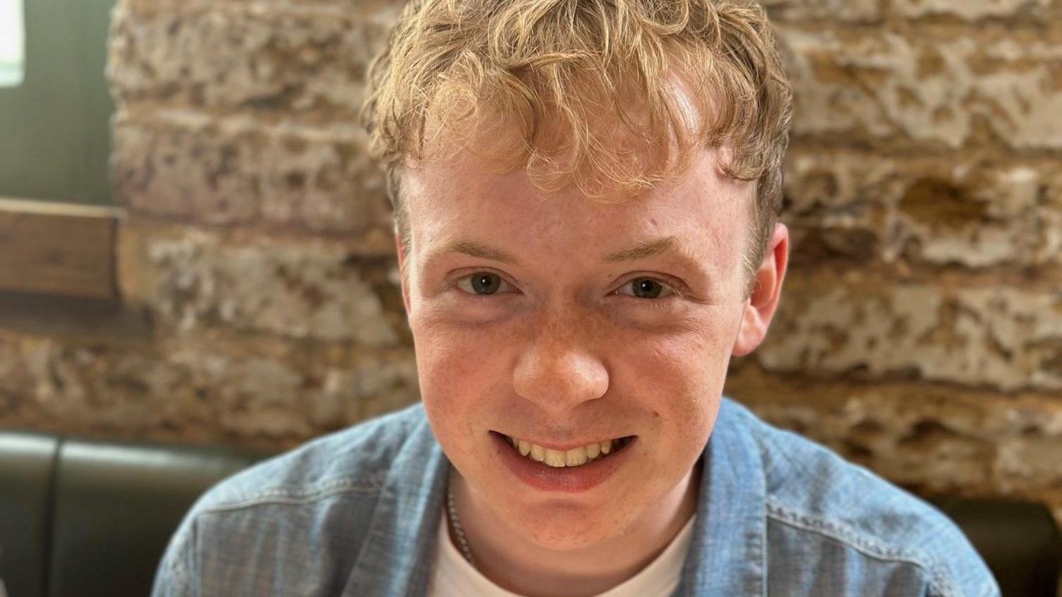 A young man with brown eyes smiles at a camera while wearing a blue denim shirt