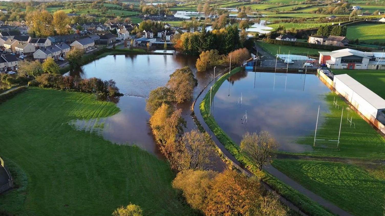 An aerial drone shot shows a small town battling flood water. The water has collected into small lakes. In the centre of the shot are two fields. The one on the right has Gaelic football posts on it and is partially covered in brown water. The field to the left is fully covered in brown water. 