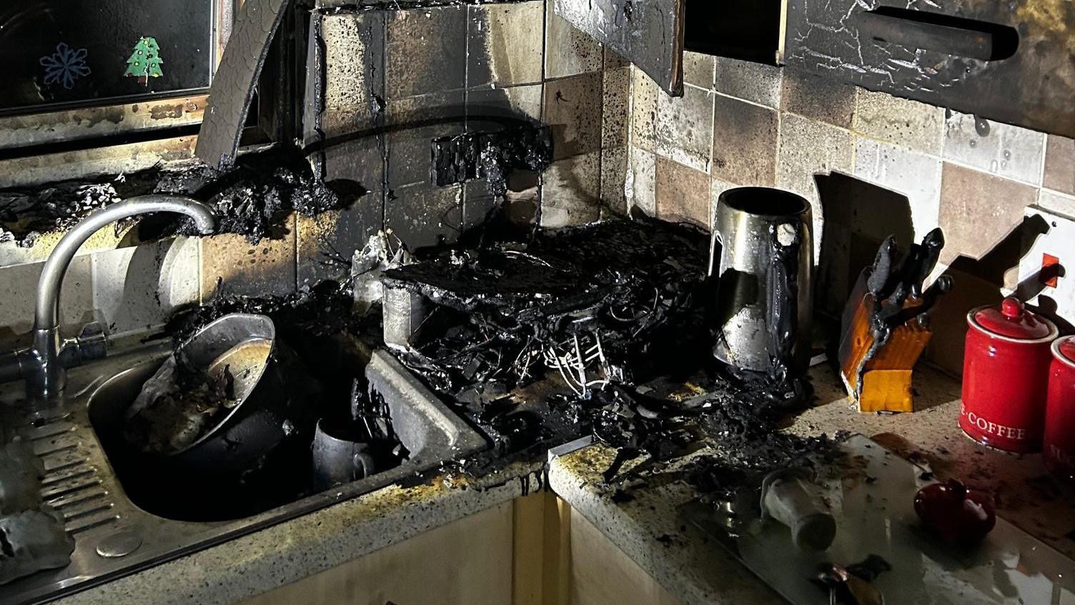 The corner of a kitchen after a fire. The cupboards are scorched black and the surfaces and sink are covered in ash.