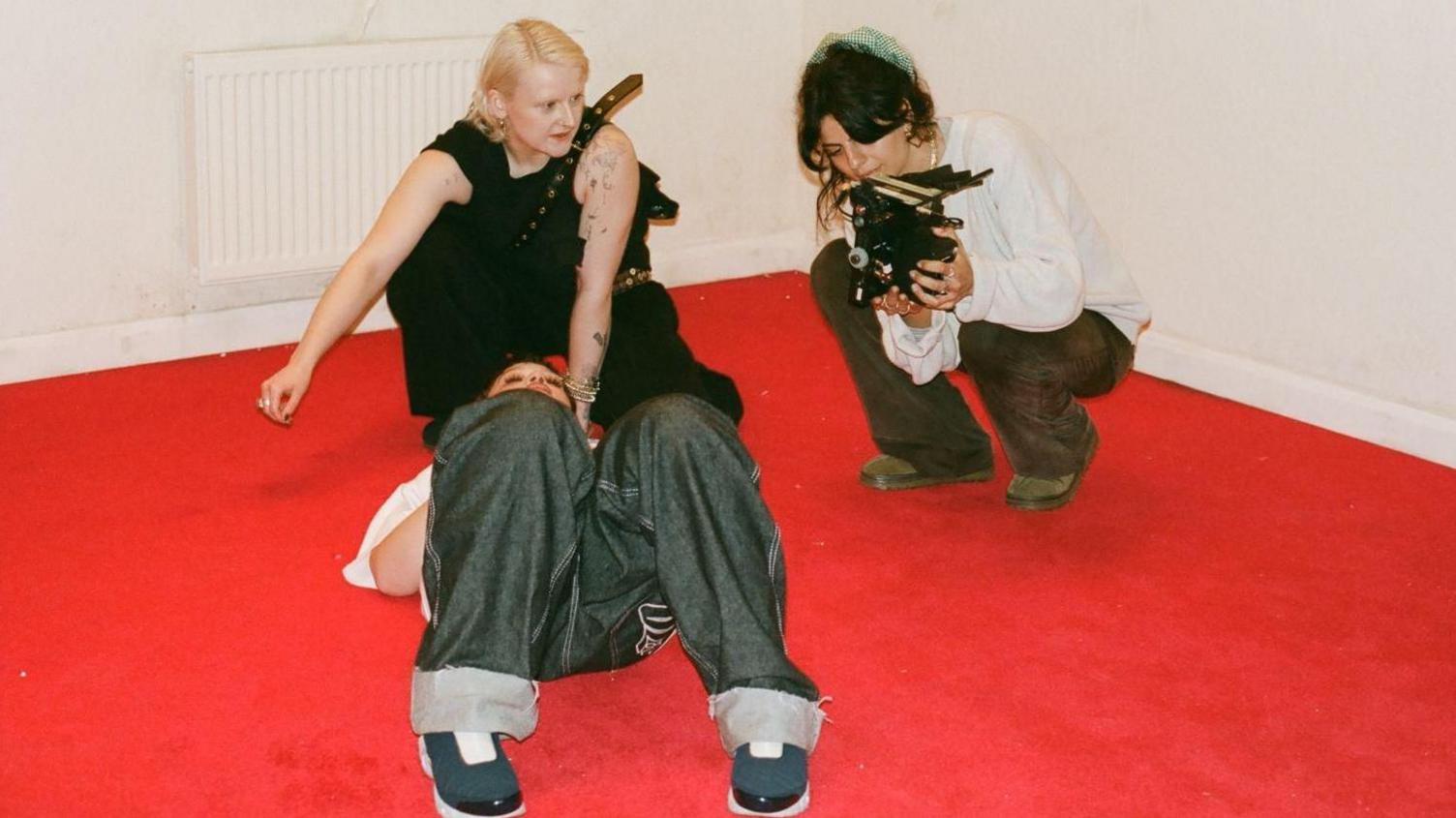 Sarah on set with Lola Young. Lola lies on her back on the red carpet with her knees up while Sarah crouches holding a camera. A third woman is there, she crouches behind Lola's head and looks at what Sarah's showing on the camera. 