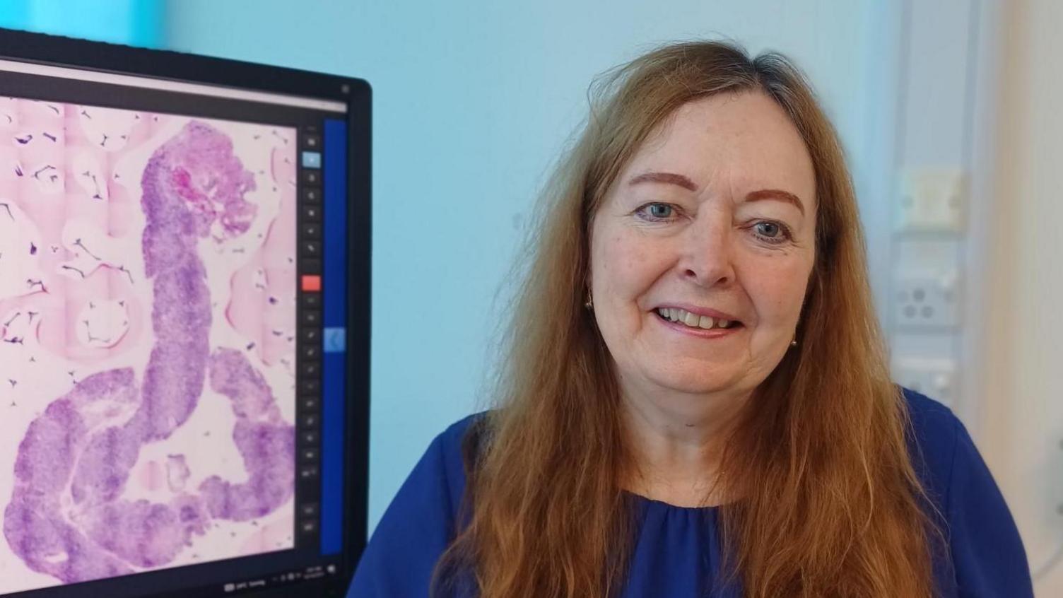 Dr Anne Warren is sitting in an office with a computer screen to her right. She has long auburn hair and is wearing a navy blue top