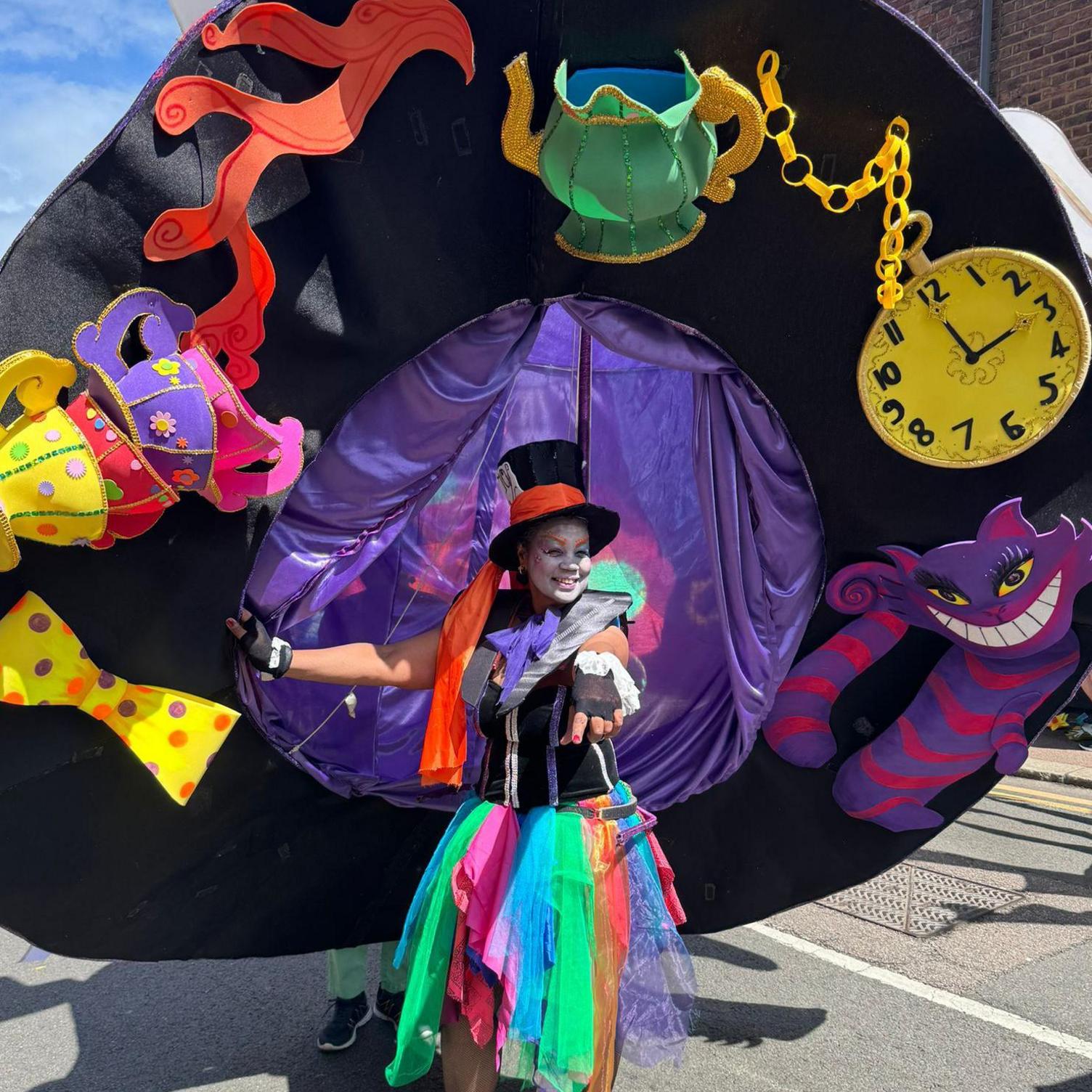 Maureen Drummond wearing an enormous hat costume which is three times her size, decorated with Alice in Wonderland characters and objects