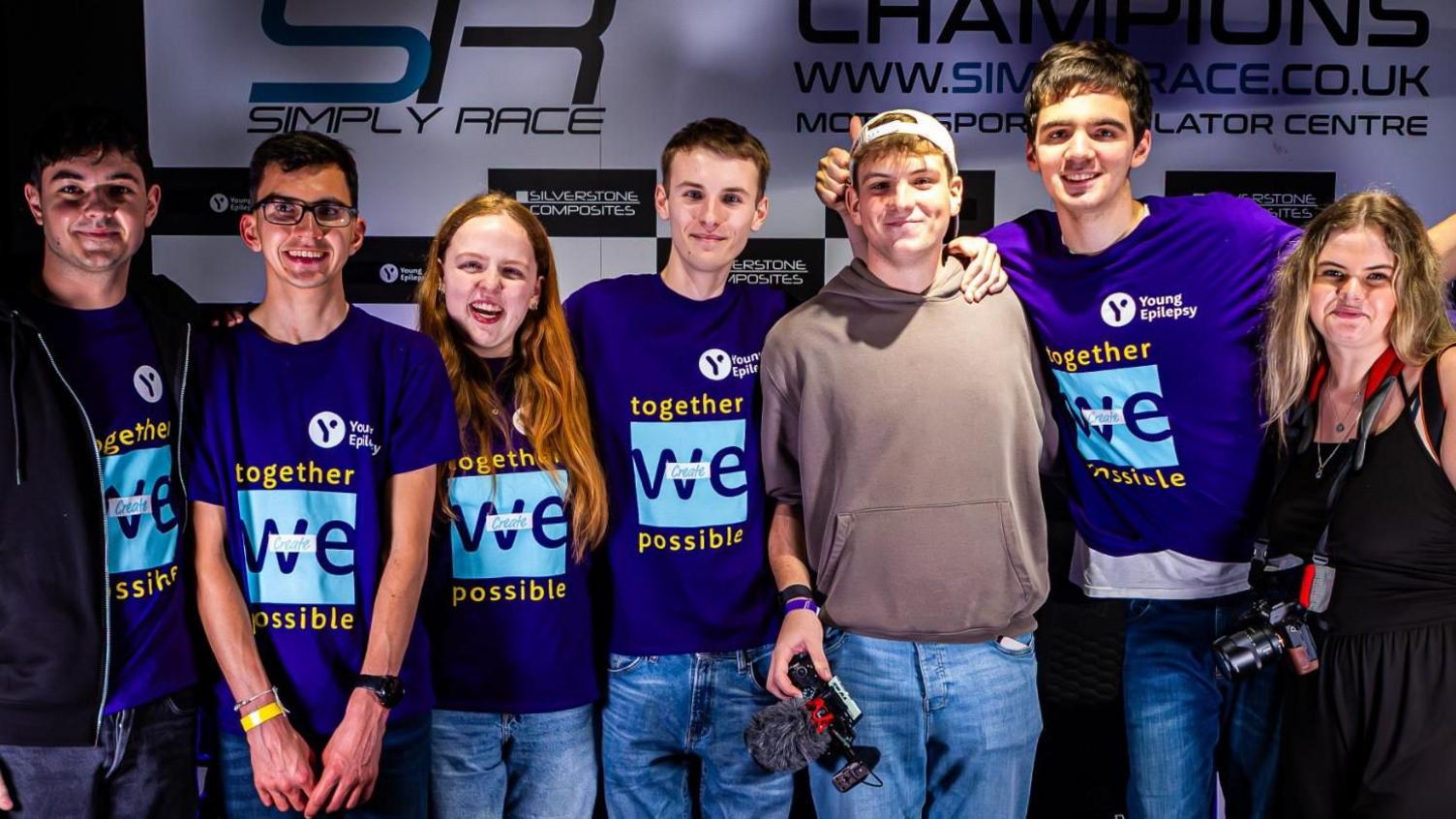 A group of seven younger people male and female all wearing purple t-shirts with blue branding in the middle. They are stood in front of a black and white advertising board.