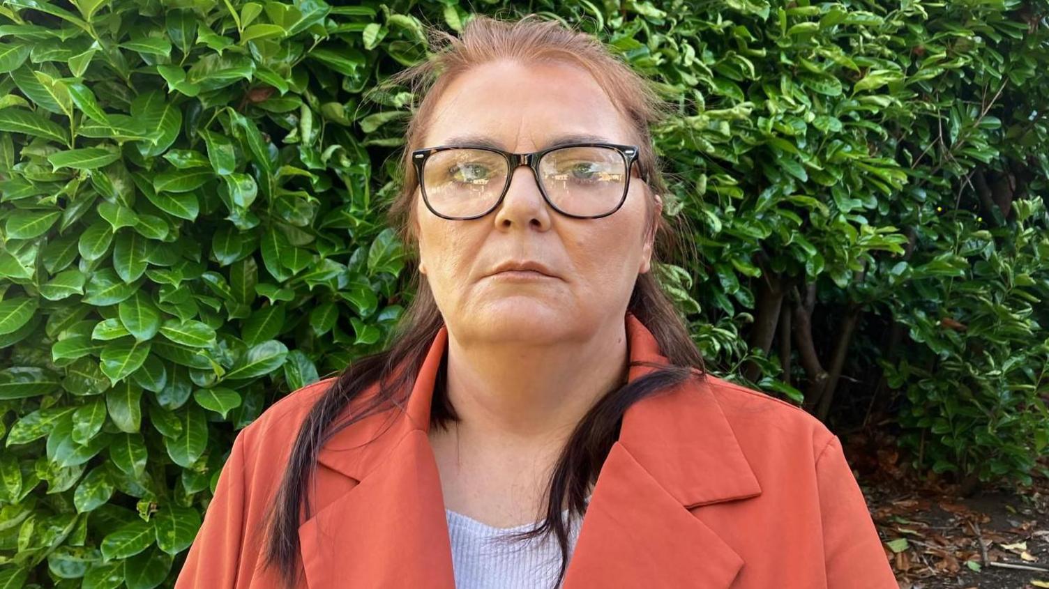 A woman with brown hair and dressed in an orange coat, wearing back glasses, staring directly at the camera. 