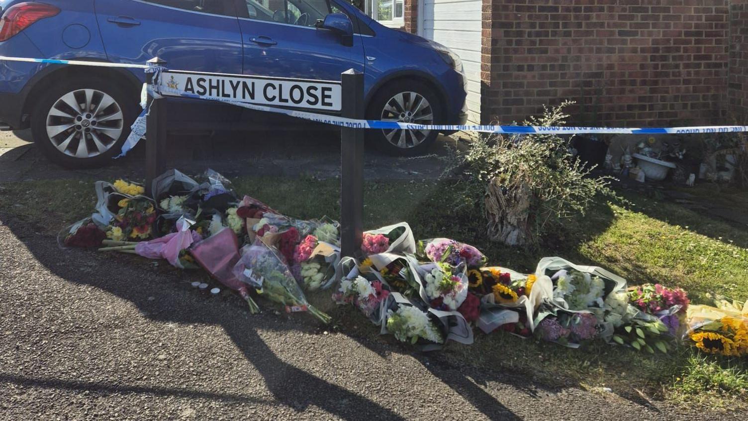 Dozens of bunches of flowers placed on a pavement, next to the street sign for Ashlyn Close in Bushey. Police cordon tape is attached to the left post of the sign and a blue car parked on a driveway is visible in the background.