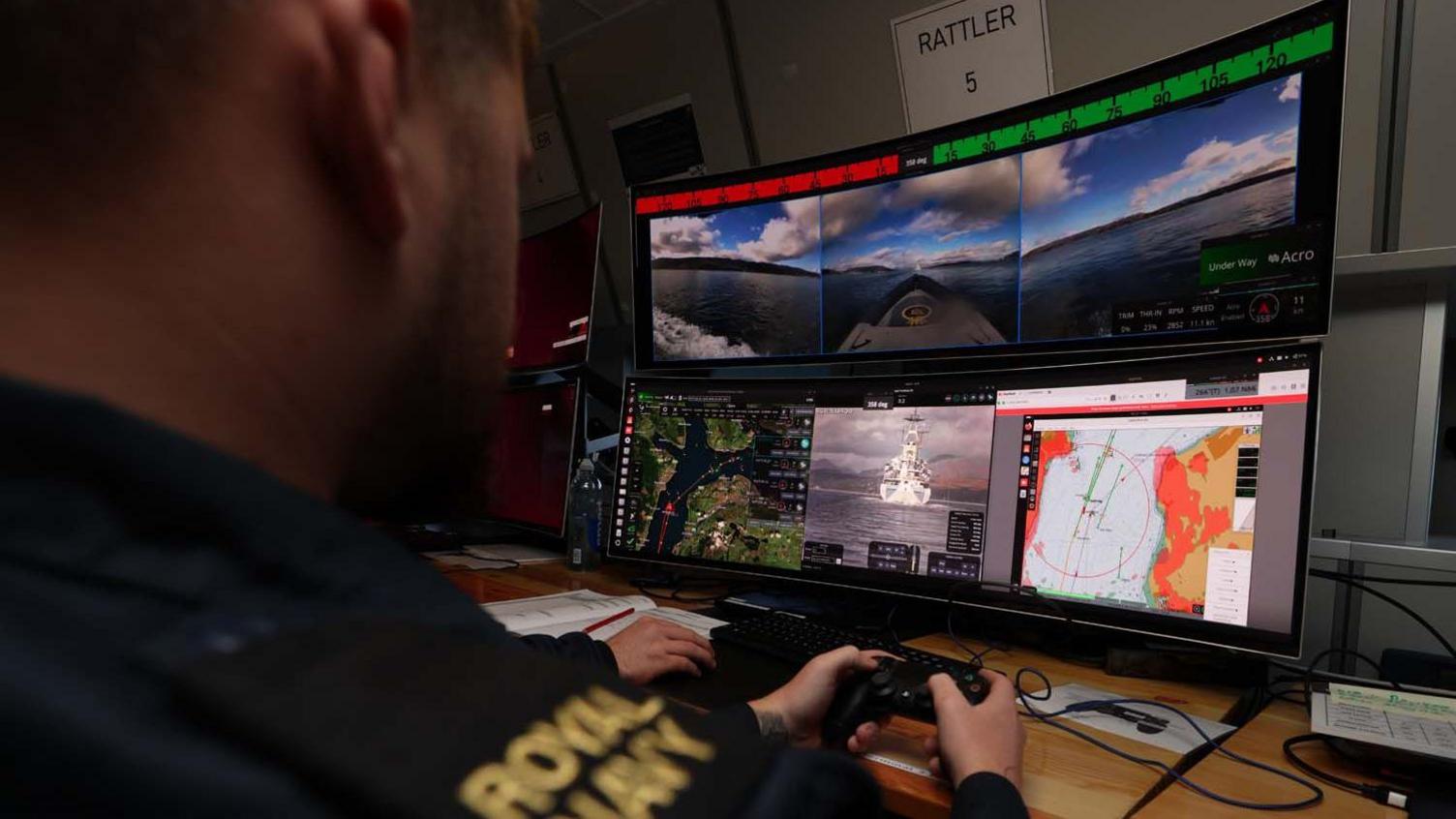 A male Royal Navy crew member sits with his back to the camera holding what looks like a black games controller and looking at a panoramic computer screen. The screen shows camera views from each boat. The man wears a dark blue shirt, which has a black panel on the shoulder and gold embroidered writing which says 'Royal Navy'.
