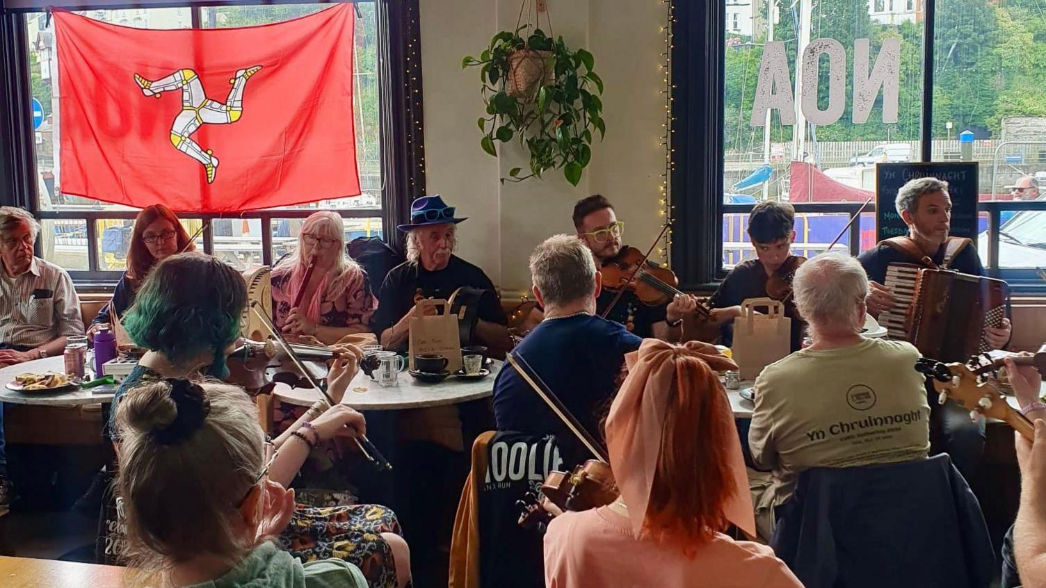 A group of musicians playing in a room. There is a Manx flag hanging in front of one of the windows.