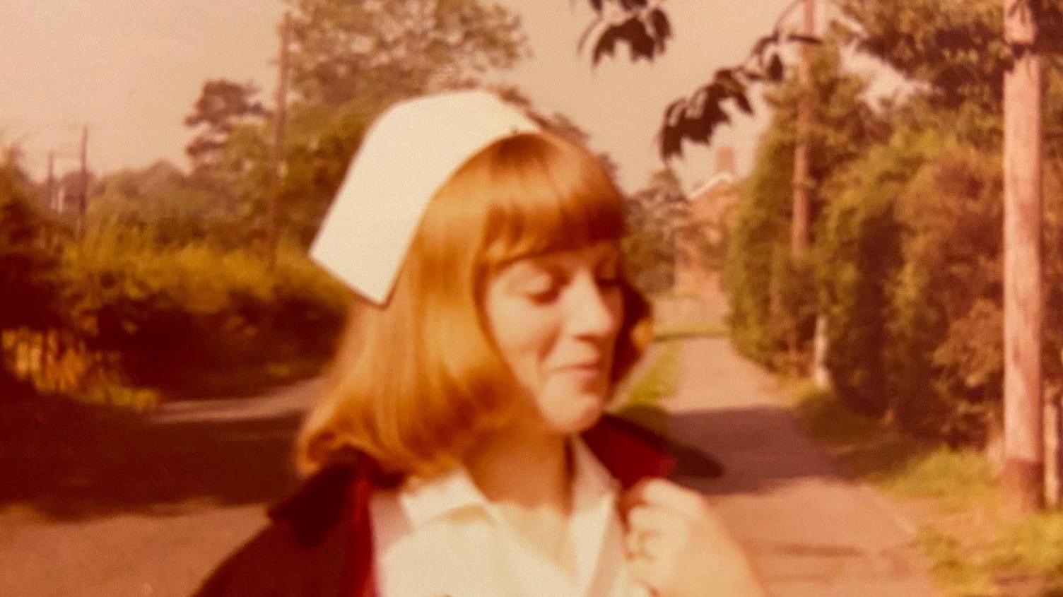 Beverley Pedley is wearing a white cap, with red hair beneath, and a white dress as well as a black cape with red lining. She is stood on a pavement lined with trees.