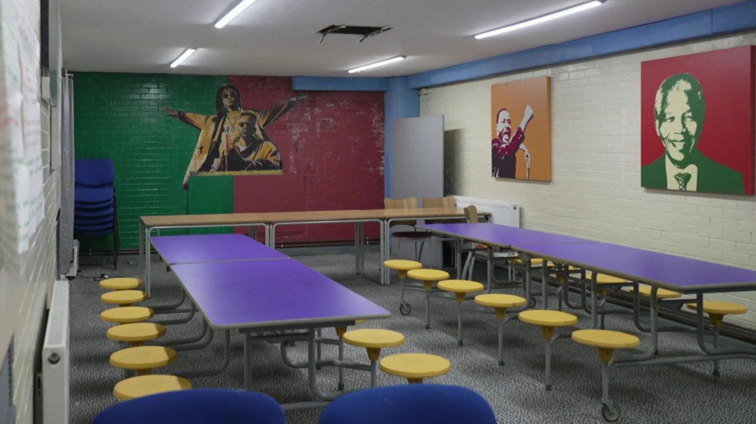 A classroom with purple tables and yellow stools and brick walls painted red, green and white with paintings of Martin Luther King Jr and Nelson Mandela.