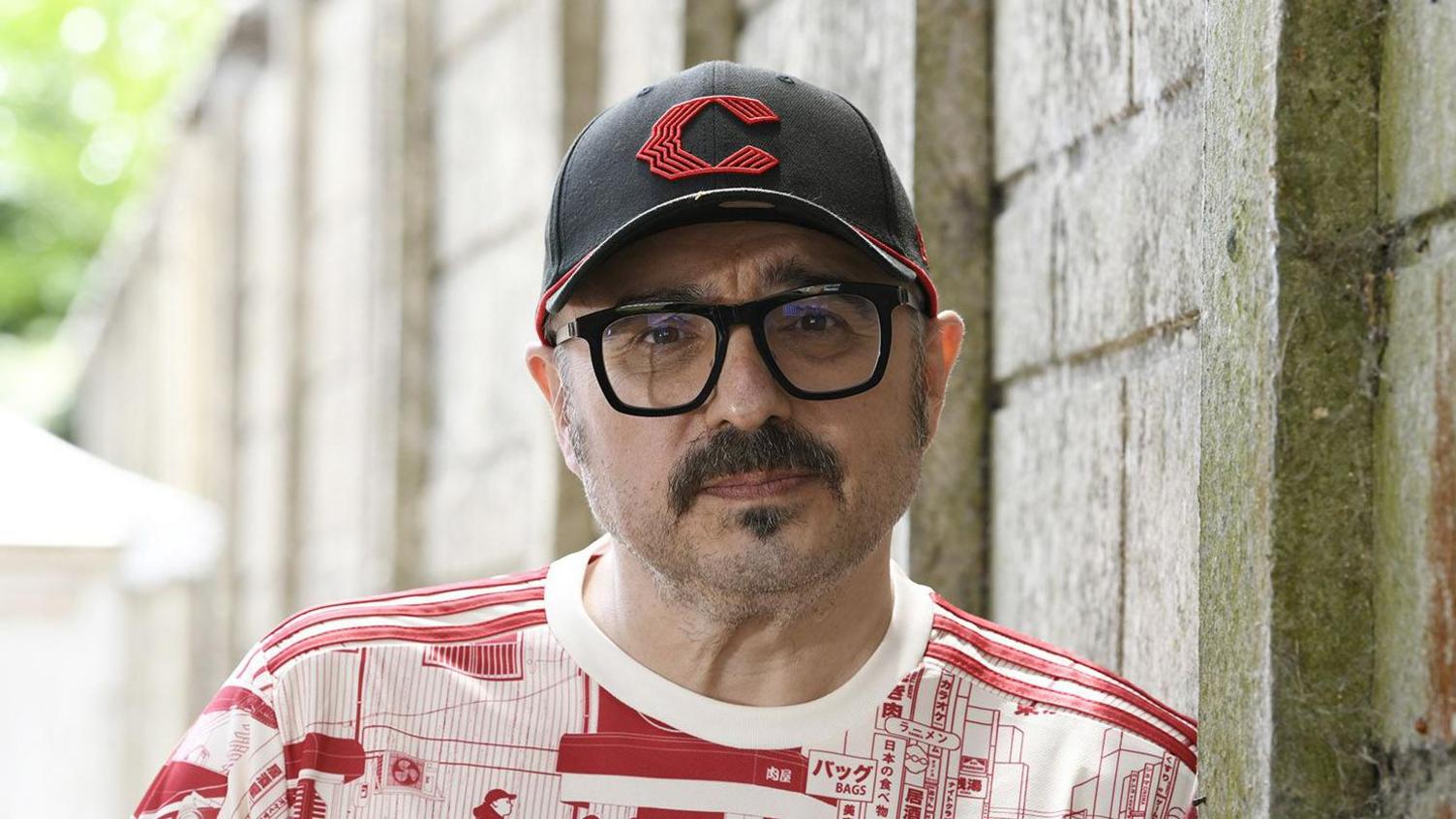 Matt Eccles has a dark moustache and is wearing a black cap, black framed glasses and a red and white T-shirt. He is leaning against an outside wall.