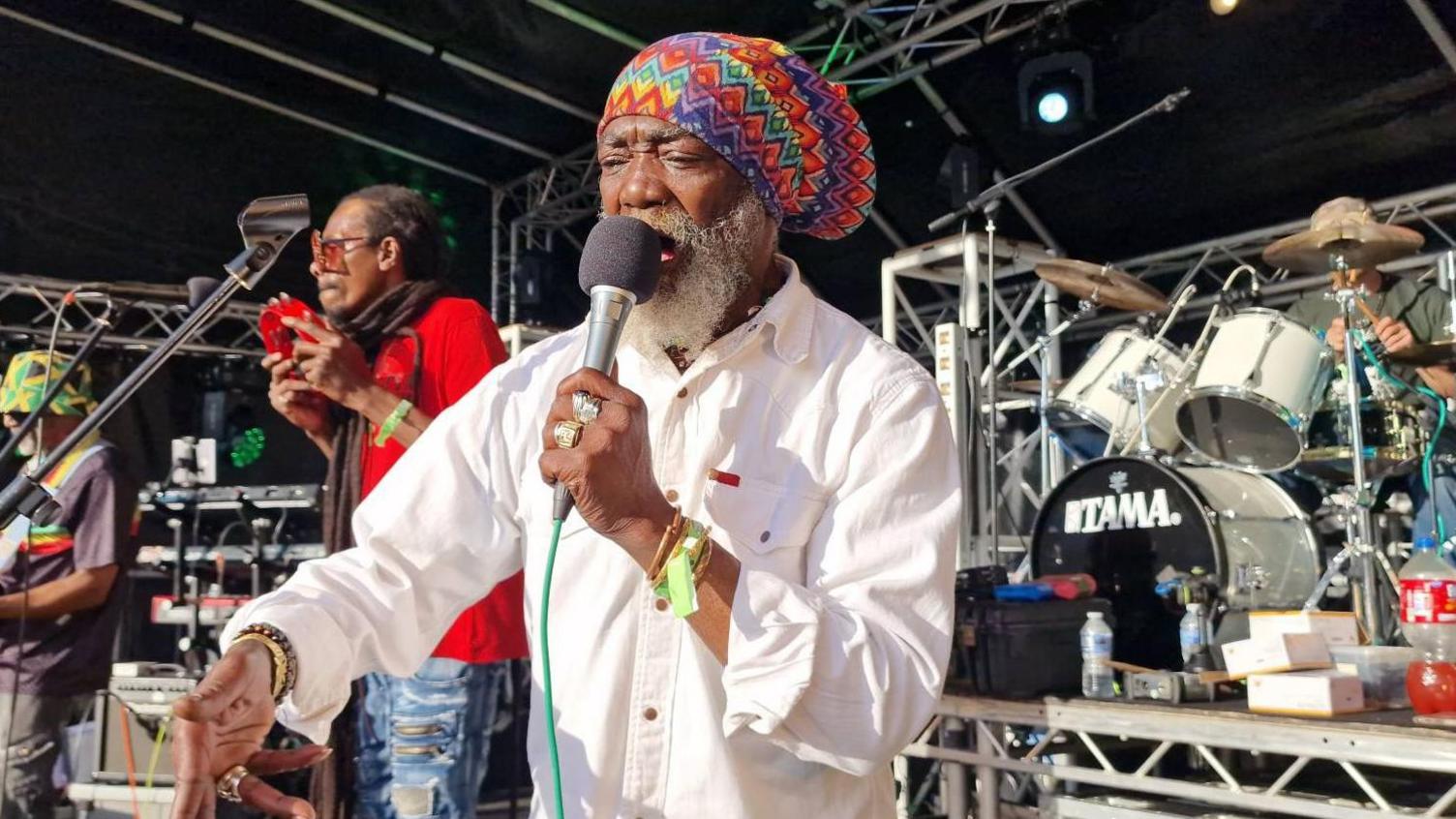 A man is wearing a multicoloured rasta cap and a white shirt. He is signing into a microphone on a stage. In the background, a man is playing the tamborine and another is playing the drums.