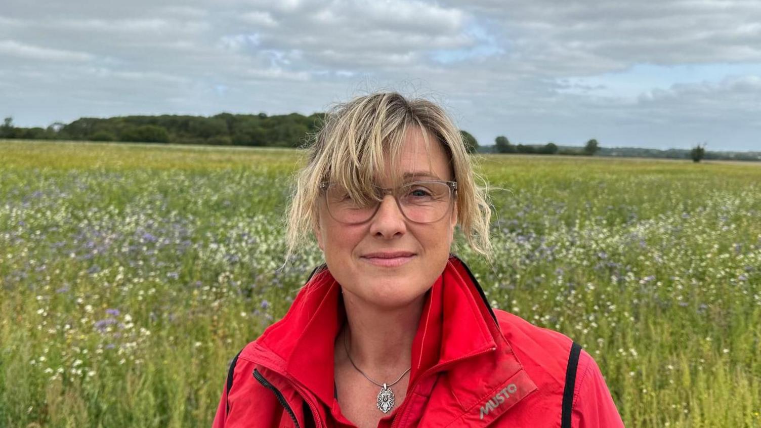 Tammie McNeill standing in front of a field, she is wearing a red jacket and glasses. She has blonde hair. The field is large with purple and white flowers.
