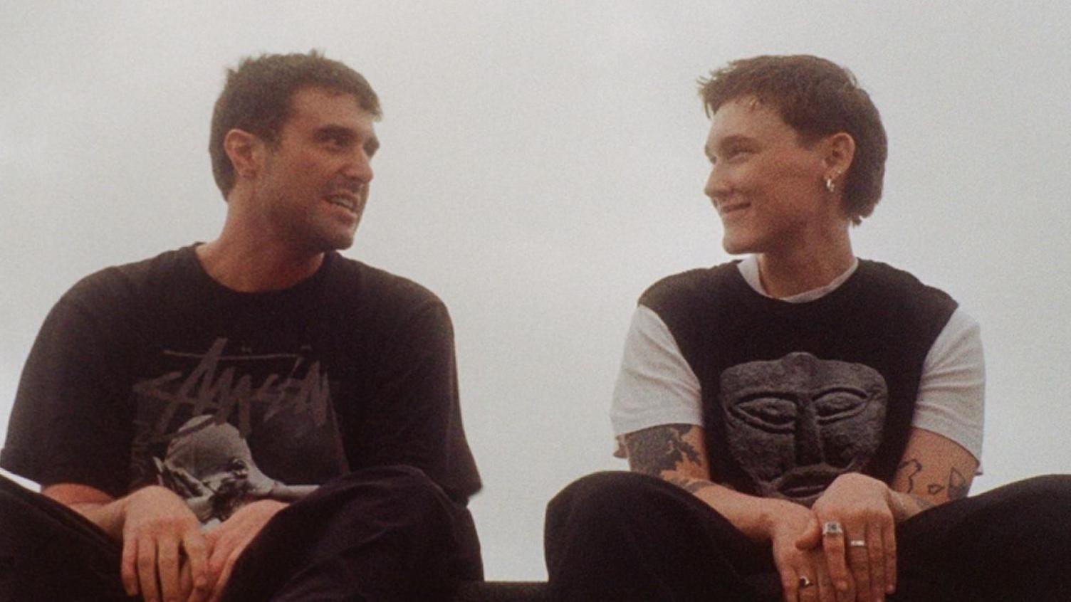 Fred Again and SOAK, both wearing black t-shirts and trousers, sitting on a ledge with grey sky in background