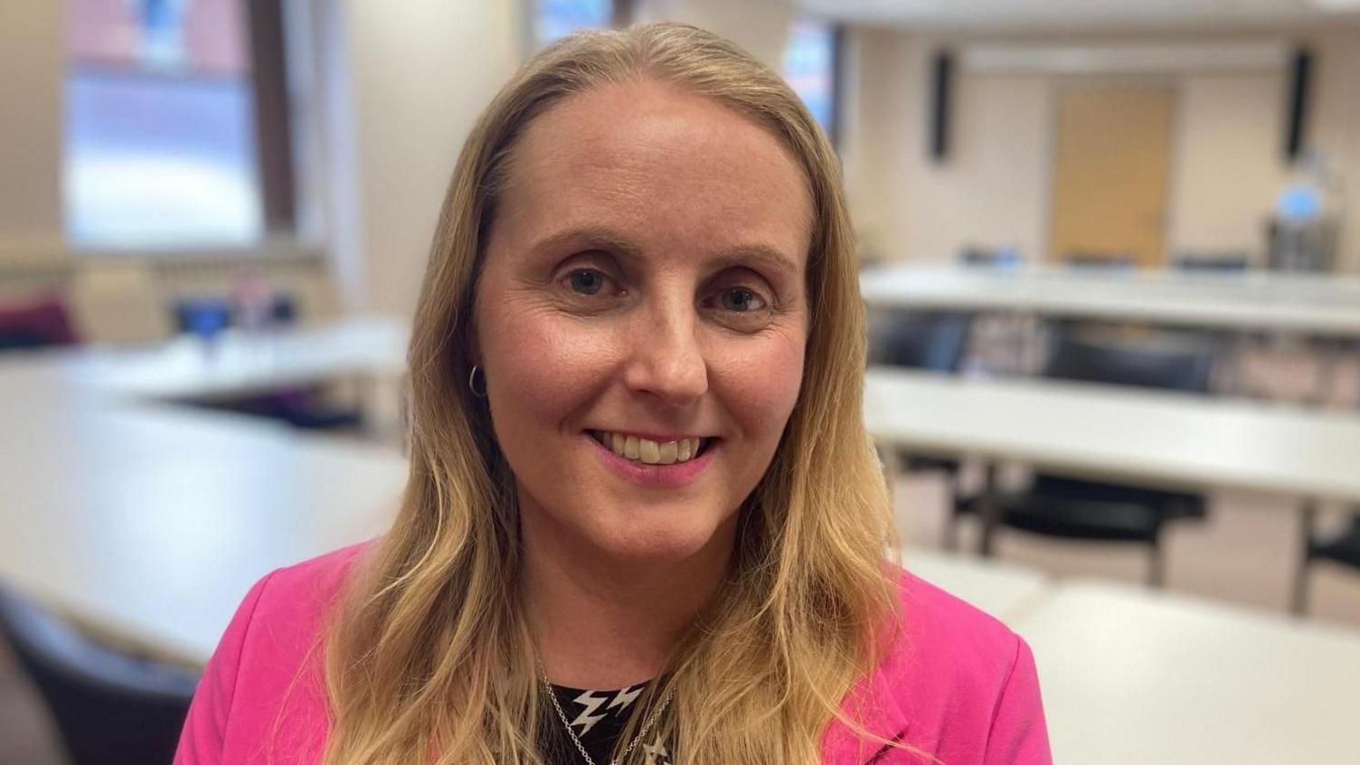 A head and shoulders photograph of councillor Naomi Tweddle. She is standing in a room with table and chairs behind her and is wearing a bright pink suit jacket.